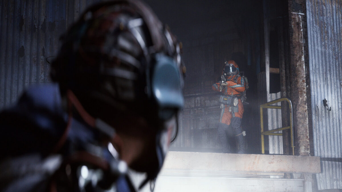 Ein Mann mit Helm und Headset, schaut zu einem anderen Mann mit Rüstung und Waffe.