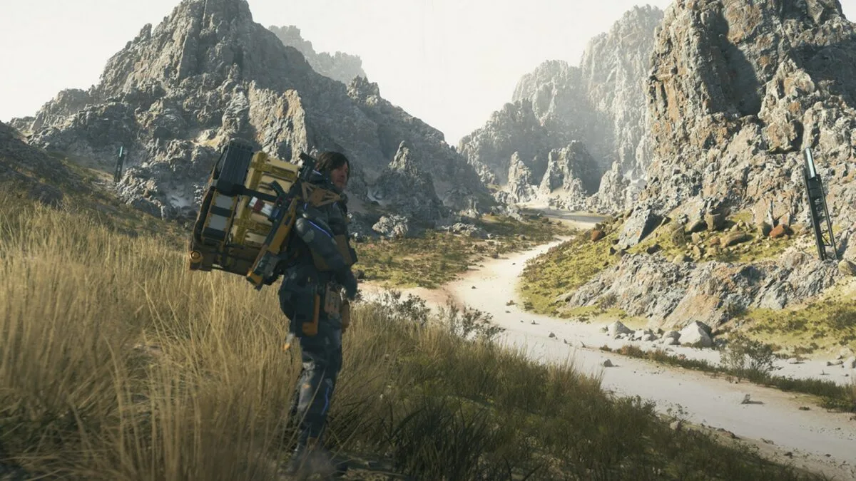 Ein Mann mit großem Rucksack steht vor einer Bergkulisse im Gras.