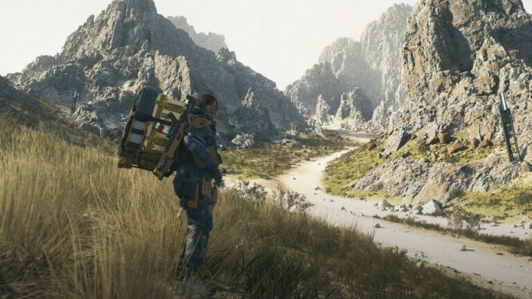 Ein Mann mit großem Rucksack steht vor einer Bergkulisse im Gras.