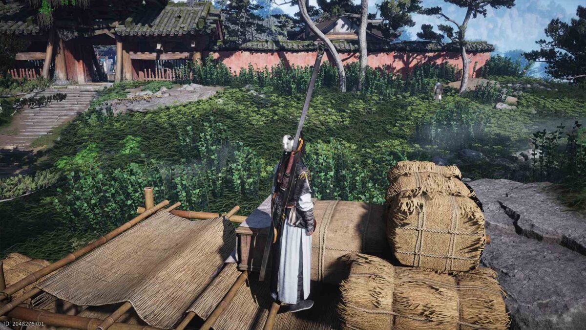 Ein maskierter Krieger in langer, dunkler Gewandung steht auf einer hölzernen Plattform neben gestapelten Strohballen. Er hält einen langen Speer über der Schulter. Im Hintergrund liegen überwucherte Steinpfade, dichter Bewuchs und eine traditionelle asiatische Toranlage aus Holz und Lehm, teilweise von Bäumen und Sträuchern umrahmt.