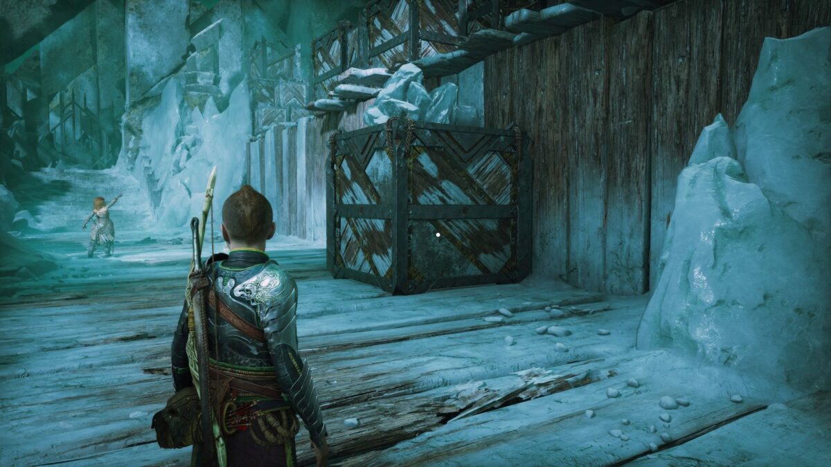 Atreus steht in God of War Ragnarök vor einem großen Holzklotz, während Thrúd auf eine Holzwand zeigt.