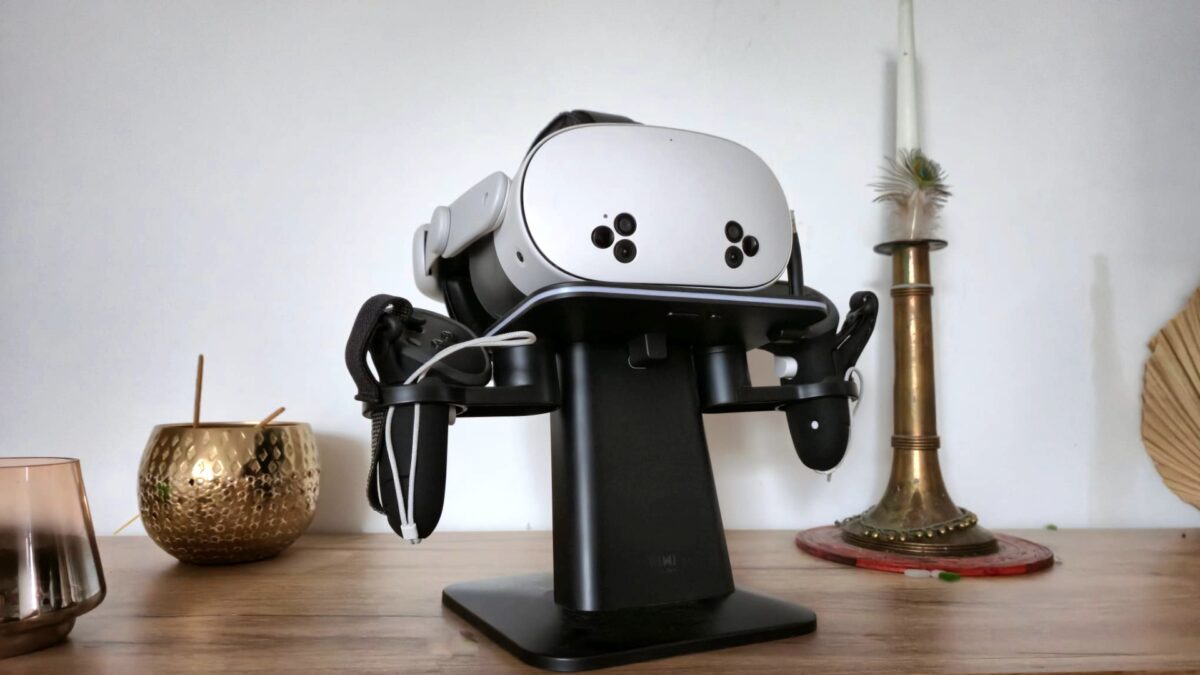 A white VR headset (Meta Quest 3S) sits on a black stand placed on a wooden table decorated with gold accents, including a candlestick and a small vase.