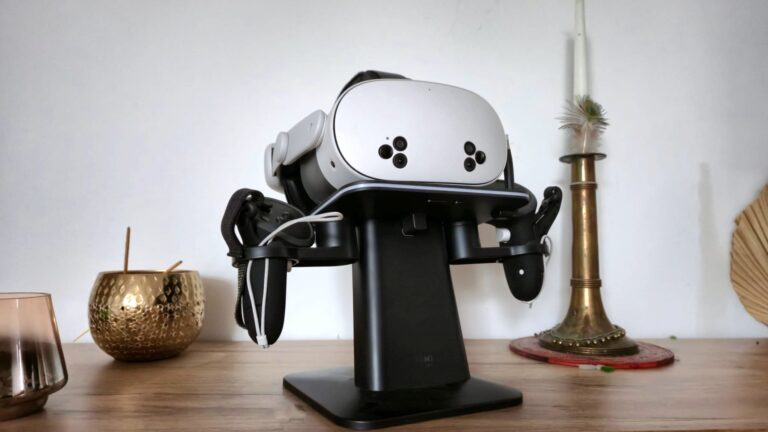 A white VR headset (Meta Quest 3S) sits on a black stand placed on a wooden table decorated with gold accents, including a candlestick and a small vase.