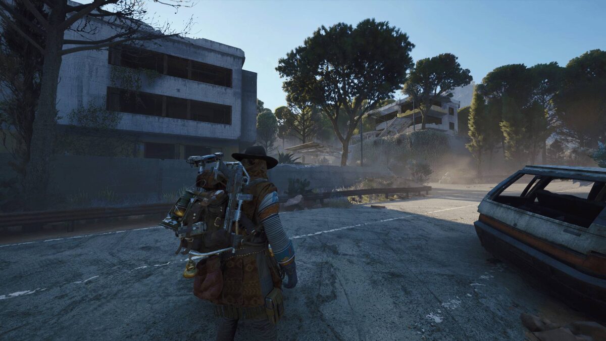 Ein Spielercharakter mit Hut und schwerem Rucksack voller Ausrüstung steht auf einer verlassenen Straße vor verfallenen Gebäuden und umgeben von Bäumen – Szene aus einem postapokalyptischen Shooter-Spiel.