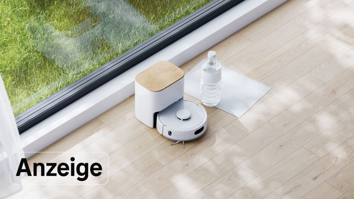 Ein Saugroboter steht in seiner Absaugstation neben einer Wasserflasche am Fenster eines Wohnzimmers.