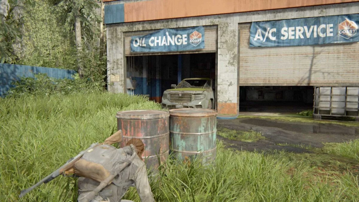 Ellie schleicht in der Deckung zweier rostiger Fässer auf eine Autowerkstatt in The Last of Us Part 2 zu.