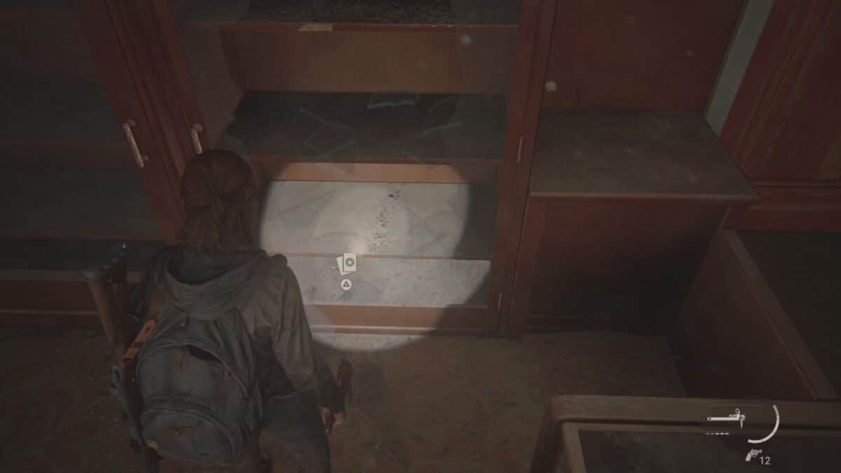 Ellie steht in The Last of Us Part 2 vor einem Schrank, dessen Glastür sie zerbrochen hat, um eine Sammelkarte aufzuheben.