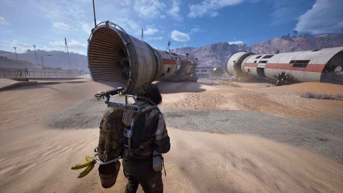 Spielcharakter steht in einer sandigen Wüste vor gigantischen abgestürzten Triebwerkssegmenten des Spaceports; klarer Himmel, Berge im Hintergrund und Staub, der über das Gelände zieht.